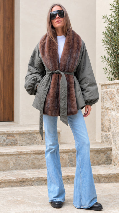 Woman wearing an olive green belted jacket with brown faux fur trim and flare jeans