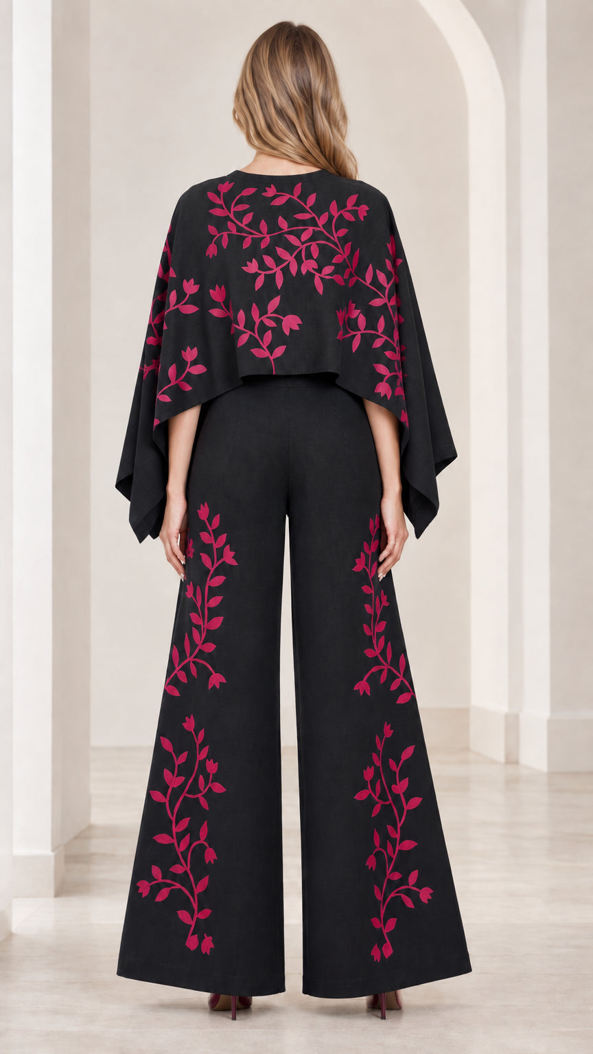 Back view of model wearing black cape wide leg jumpsuit with pink vine embroidery